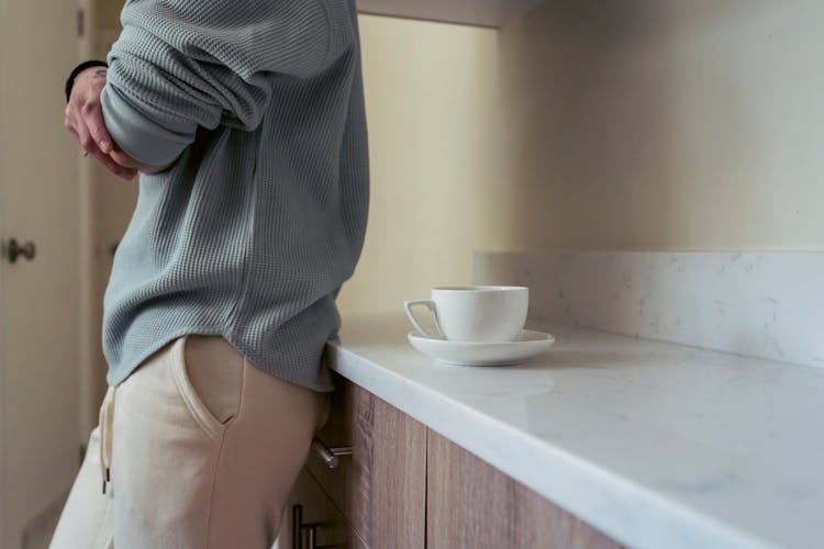 Anonymous Person Standing In Kitchen Near Cupboard With Cup Of Coffee