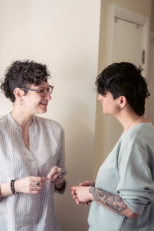 Cheerful young female best friends with dark short hair in casual outfits smiling and looking at camera while chatting at home