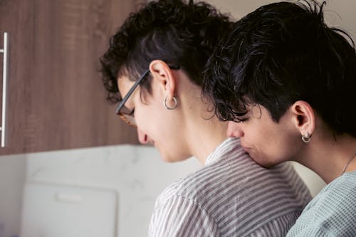 Free Side view of young homosexual androgynous woman embracing back of girlfriend with closed eyes while standing together in kitchen at home Stock Photo