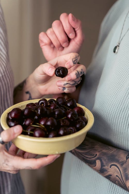 Free Crop anonymous couple with tattoos on arms in casual clothes eating bowl of appetizing ripe cherries at home Stock Photo