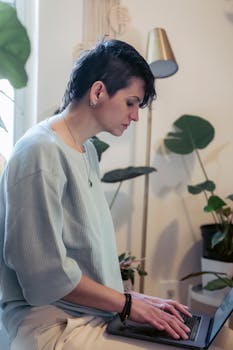 Side view of focused young woman with short hair in casual clothe working online on laptop sitting on chair in light apartment
