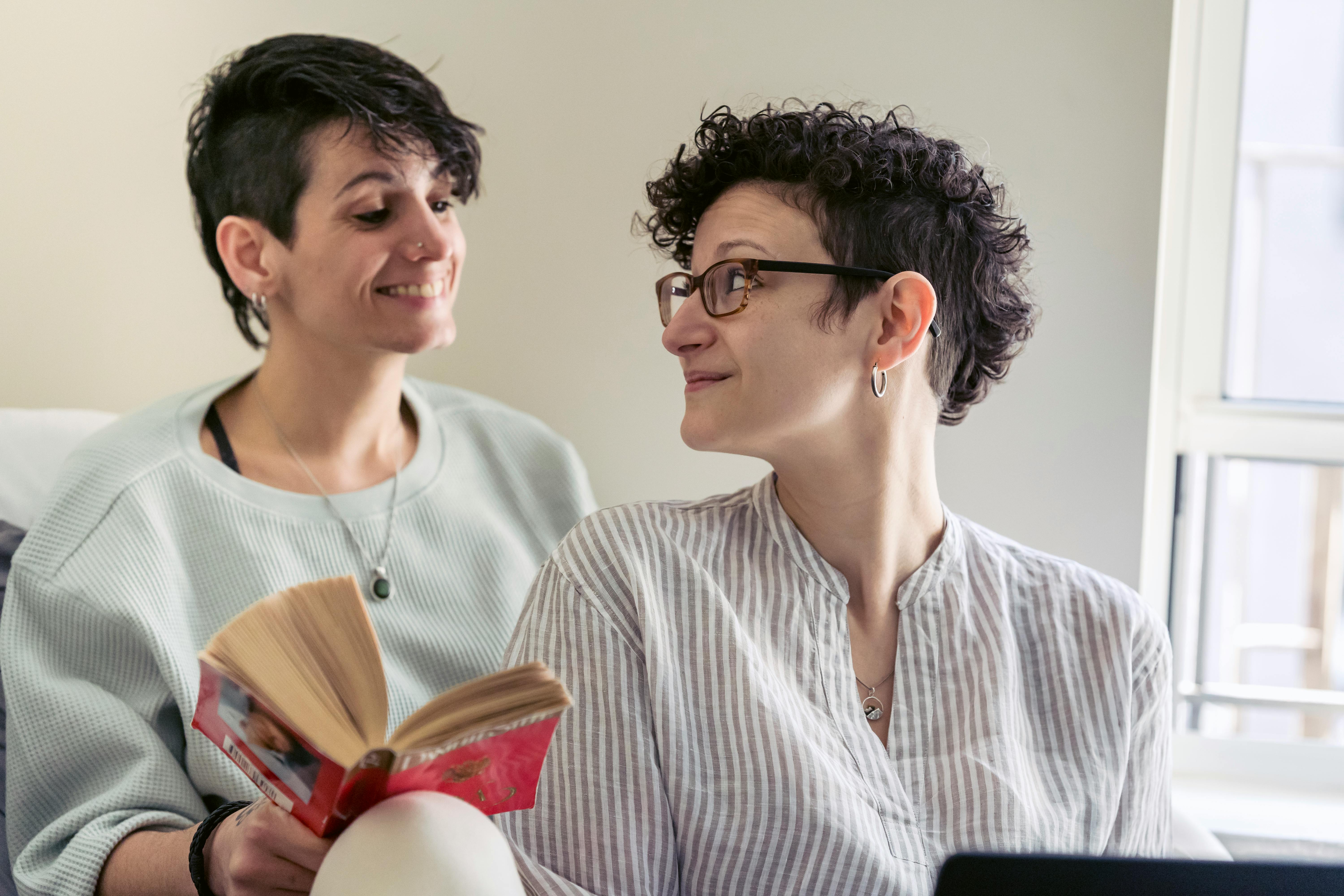 smiling lesbian couple looking at each other while reading book and using laptop