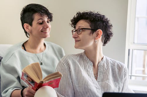 Free Cheerful young lesbian woman with short dark hair reading book while sitting behind girlfriend using laptop during weekend at home Stock Photo
