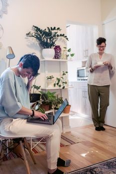 Joyful young woman with short hair working remotely on laptop at home and laughing with female best friend drinking cup of coffee