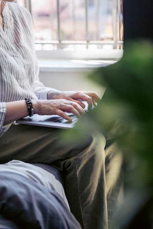 Crop faceless female freelancer in casual clothes sitting on comfortable sofa and typing on netbook while working distantly in modern apartment