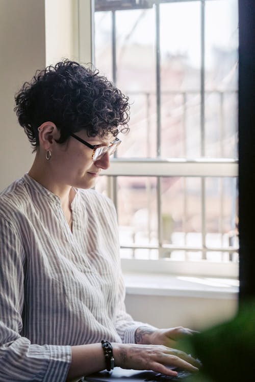 Focused female remote employee in eyeglasses and striped shirt working on netbook against window in house