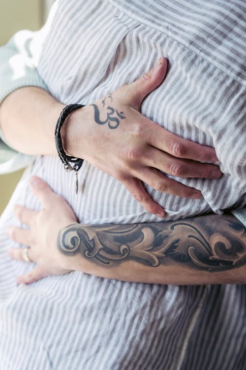 Crop anonymous wife in bracelet with ornamental tattoos embracing gently partner in striped shirt