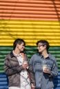 Content loving homosexual women with takeaway beverages looking at each other while standing on colorful background with LGBT rainbow flag