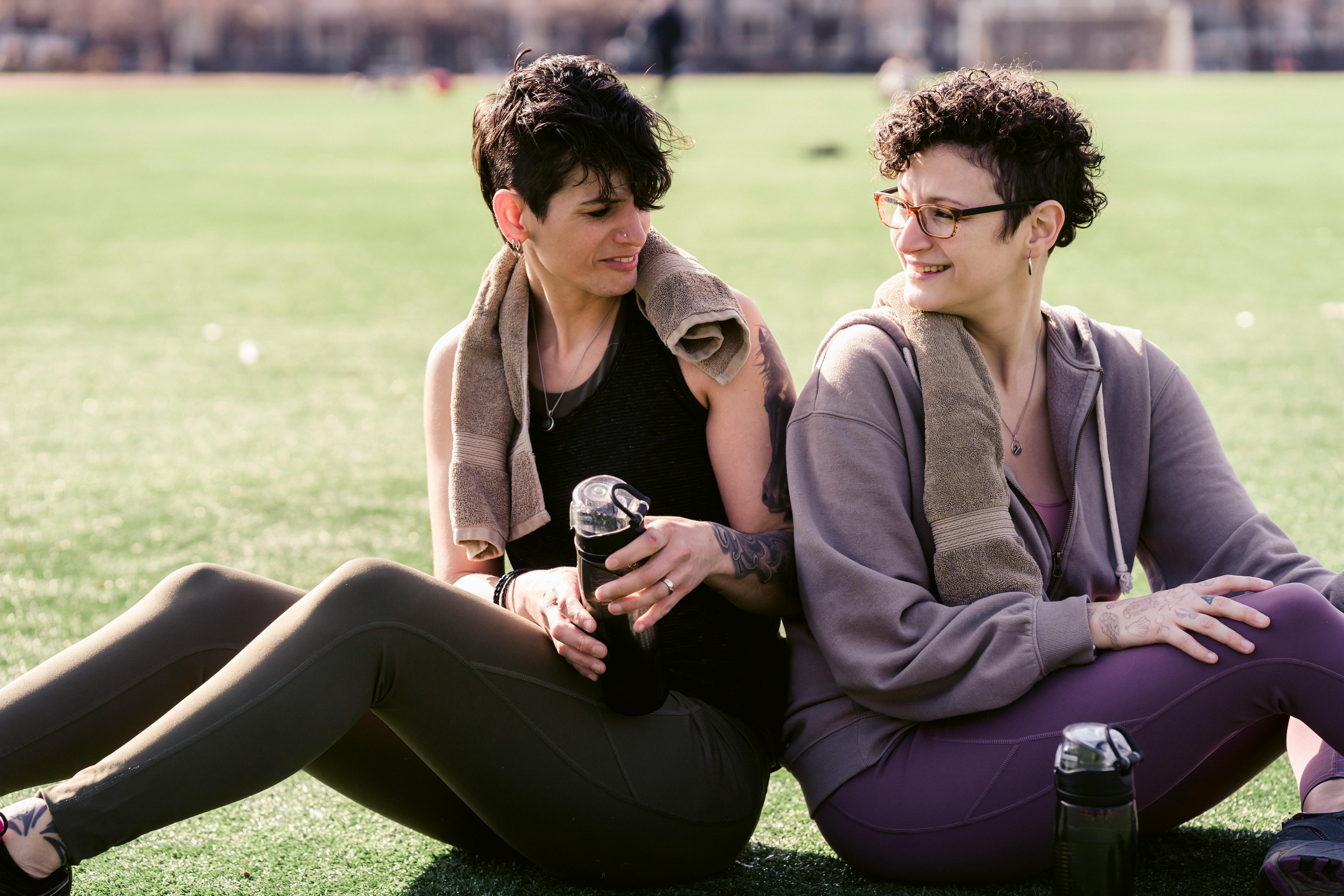 Cheerful female friends in activewear with bottles of water looking over shoulder at each other while sitting back to back on grass during training