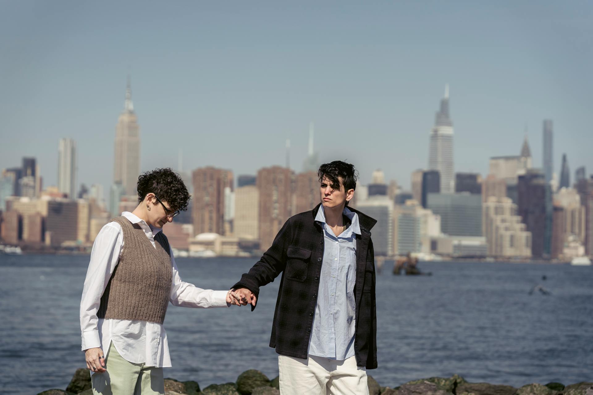 Lesbian couple holding hands, strolling by the waterfront with the New York City skyline in the background.