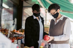 Concentrated young female friends in casual clothes and medical masks choosing fruits in local street food market on sunny day during coronavirus pandemic