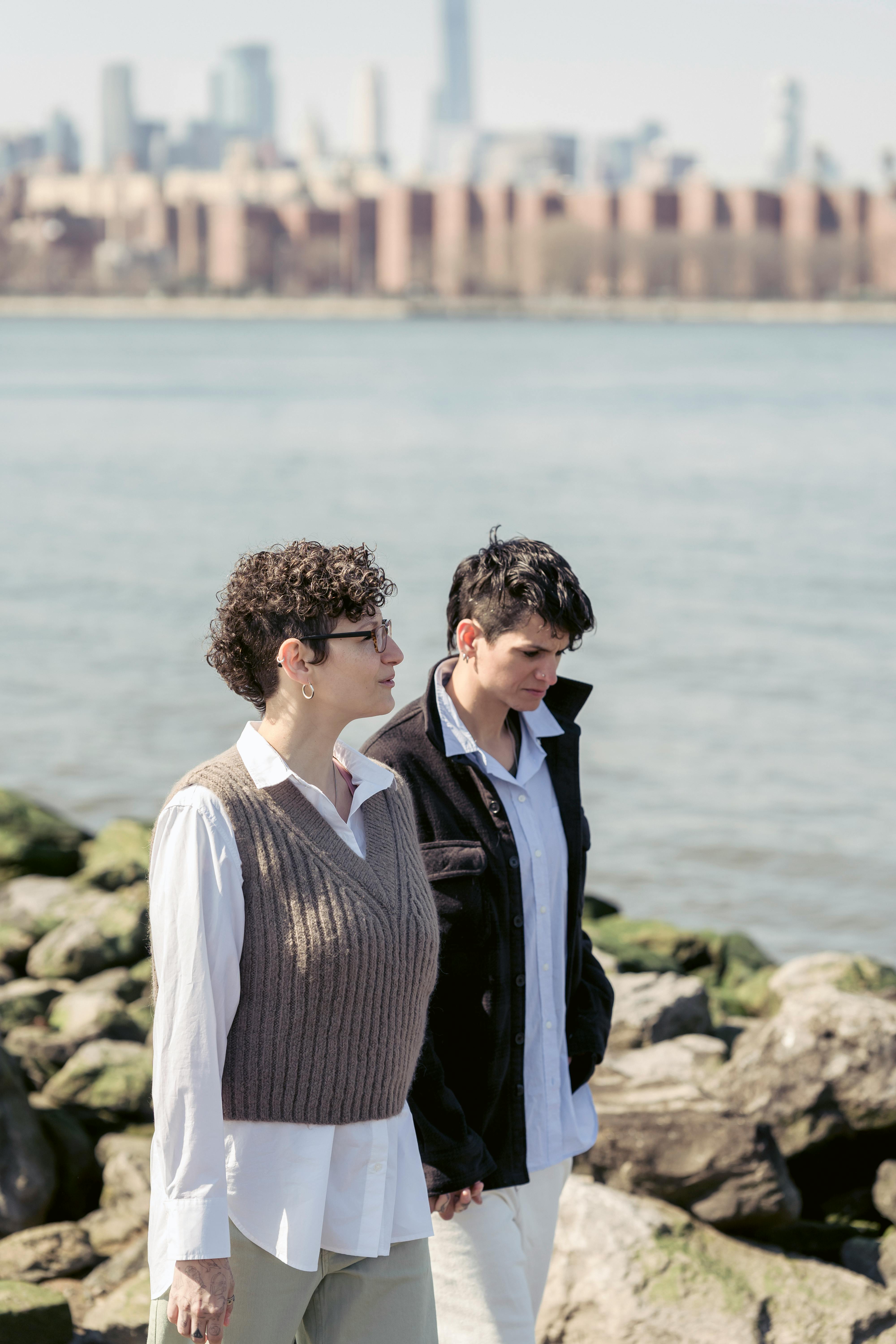 tranquil young lesbians holding hands and strolling on stony river coast in city