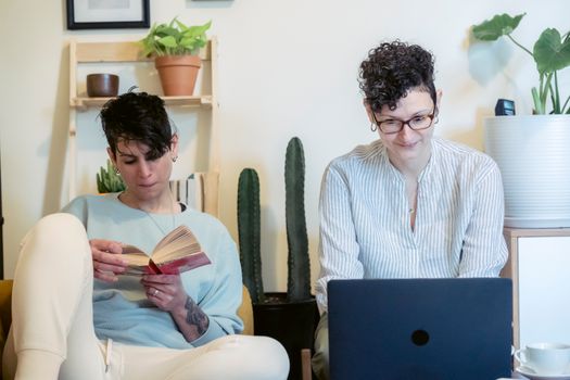 Positive young female freelancer with short dark hair in casual clothes and eyeglasses working remotely on laptop while sitting near female friend reading interesting book at home