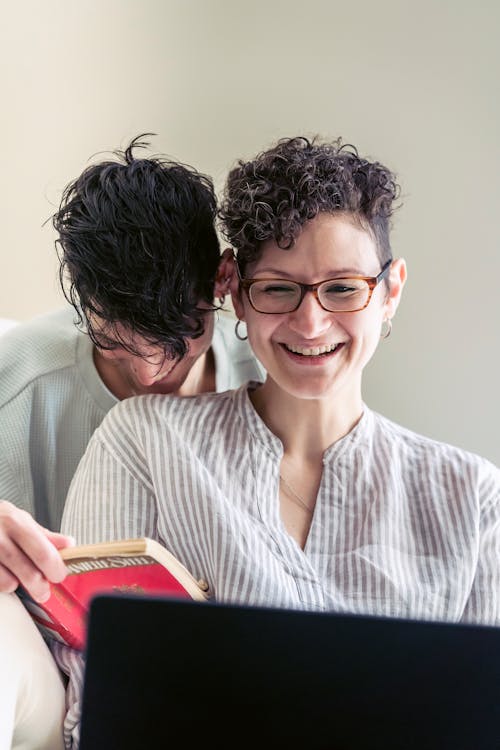 Free Laughing lesbian couple sitting on couch with book and laptop Stock Photo