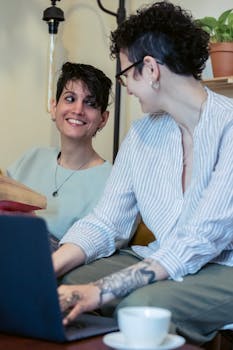 Two women with short hair smiling and interacting while using a laptop indoors.