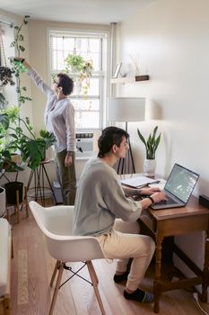 A serene home office with two adults, one working on a laptop and the other tending to plants.