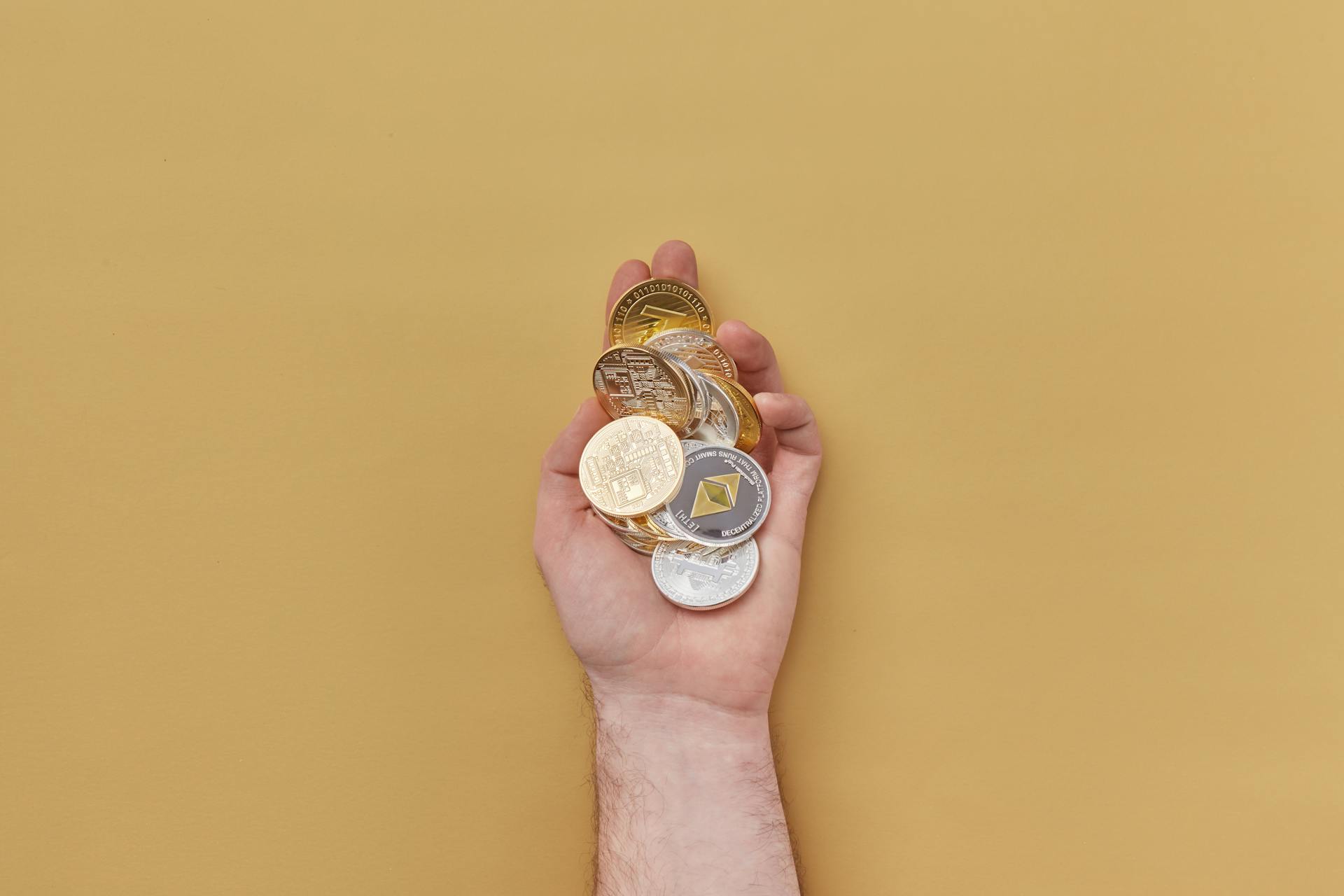 Close-up of a hand holding various cryptocurrency coins against a yellow background.