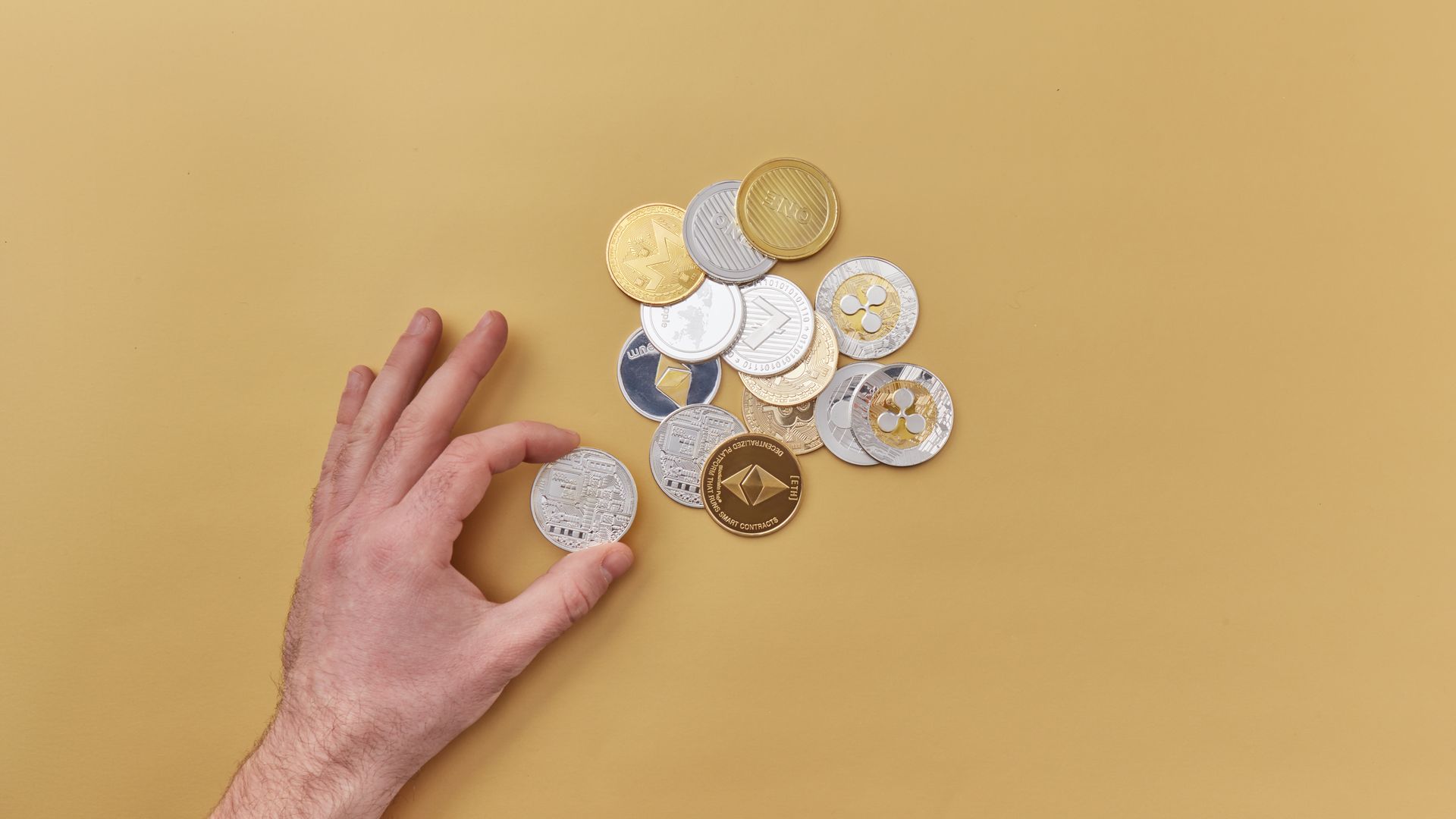 A hand picks cryptocurrencies from a mix of gold and silver coins on a vibrant yellow background — tokenizasyon