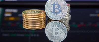 Physical Bitcoin tokens sit on a reflective surface in front of a dark screen displaying red and green financial trading charts.