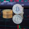 Physical Bitcoin tokens sit on a reflective surface in front of a dark screen displaying red and green financial trading charts.