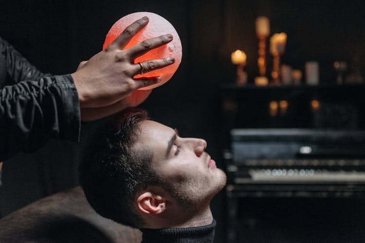 Person Holding An Illuminated Round Object On A Man's Forehead