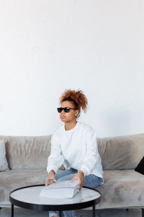 Free Image of a blind woman in sunglasses reading a Braille book on a sofa in a bright, modern living room. Stock Photo