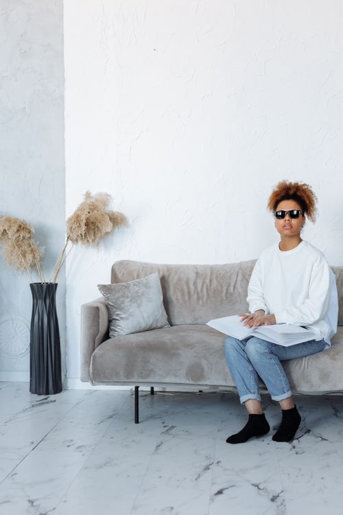 Free African American woman wearing sunglasses reads Braille book on a couch, highlighting accessibility. Stock Photo