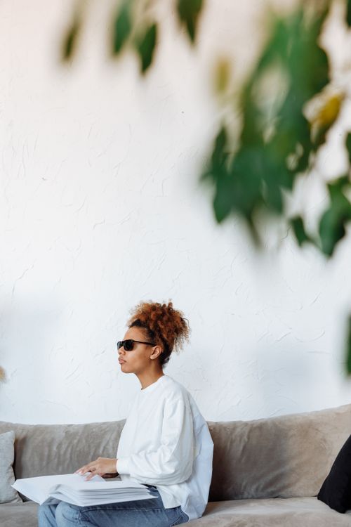 Free Woman wearing sunglasses reading a Braille book on a couch, highlighting accessibility and independence. Stock Photo