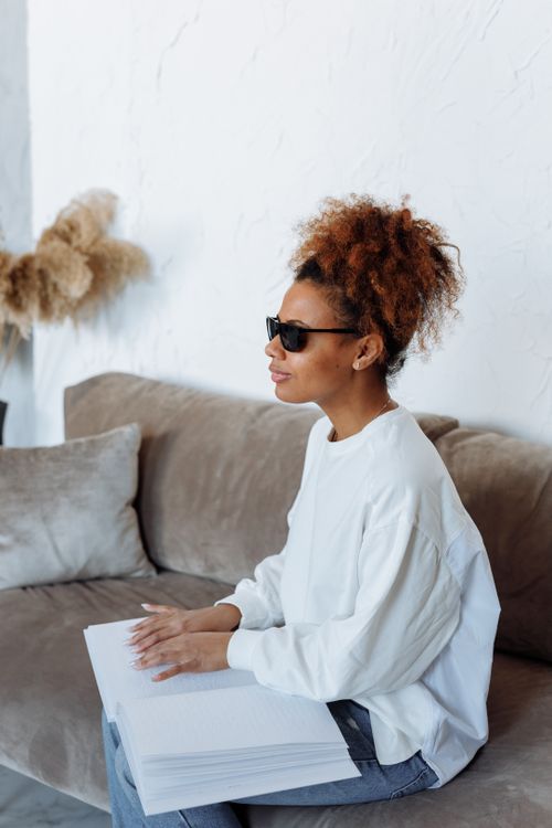Free African American woman in sunglasses reading braille book on a sofa indoors. Stock Photo