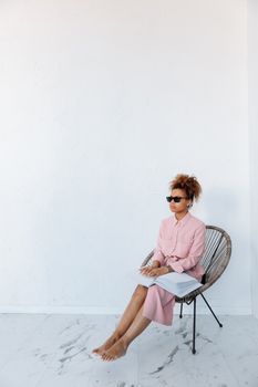 A blind woman in a pink dress reads a braille book while sitting barefoot on a chair.