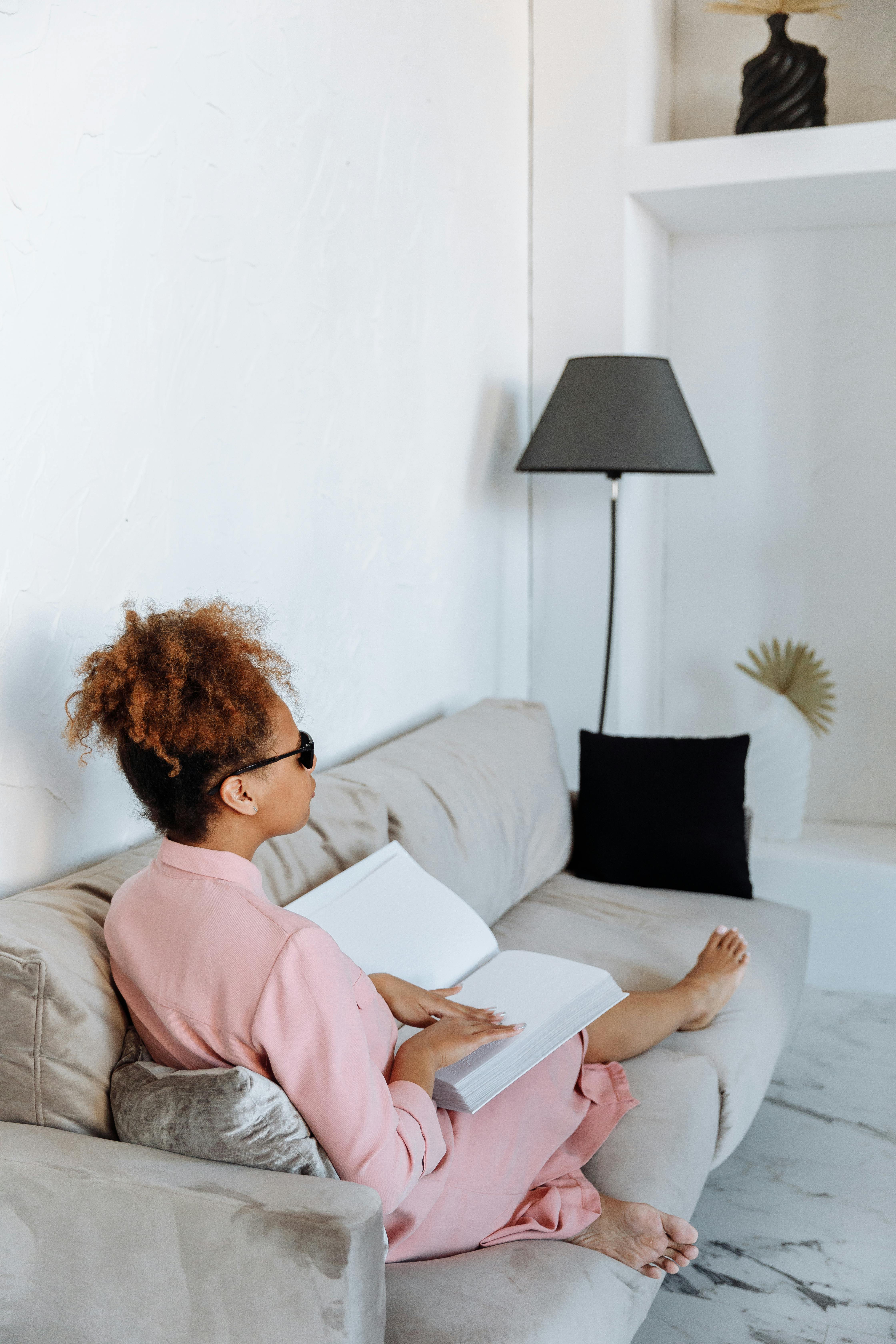 Free African American woman reading a Braille book on a couch, emphasizing disability pride and literacy. Stock Photo