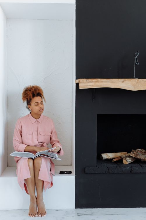 Free A woman in pink reads a Braille book by a modern fireplace, highlighting disability awareness. Stock Photo