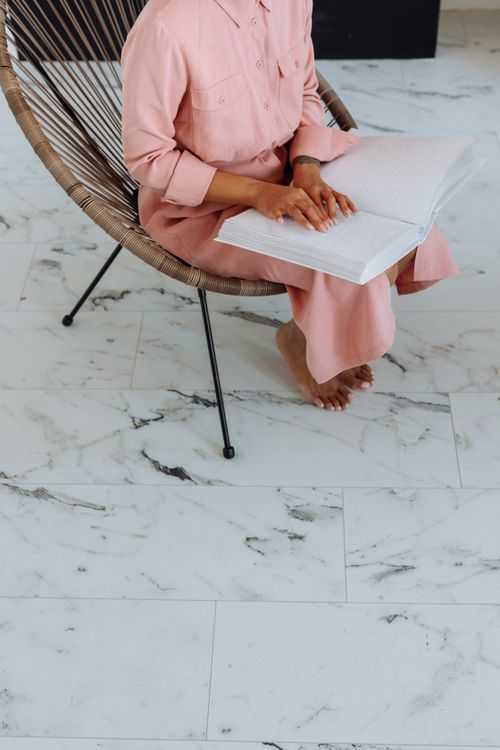 Free Woman in pink dress reading a Braille book while sitting on a woven chair indoors. Stock Photo