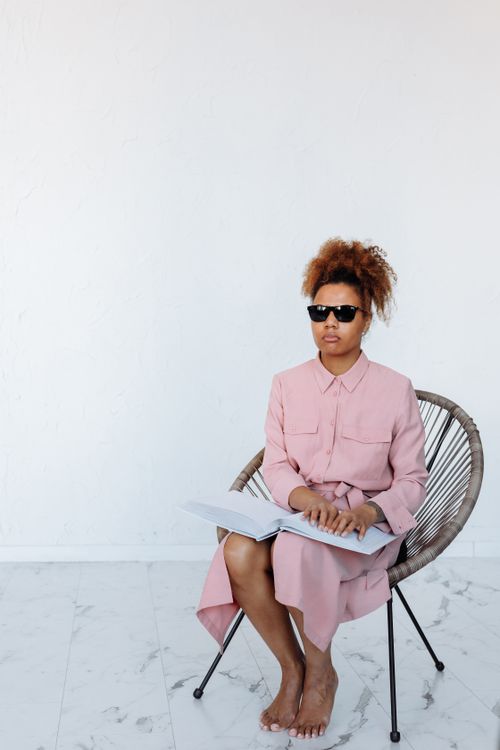Free A woman wearing sunglasses reads a Braille book while sitting indoors on a chair. Stock Photo