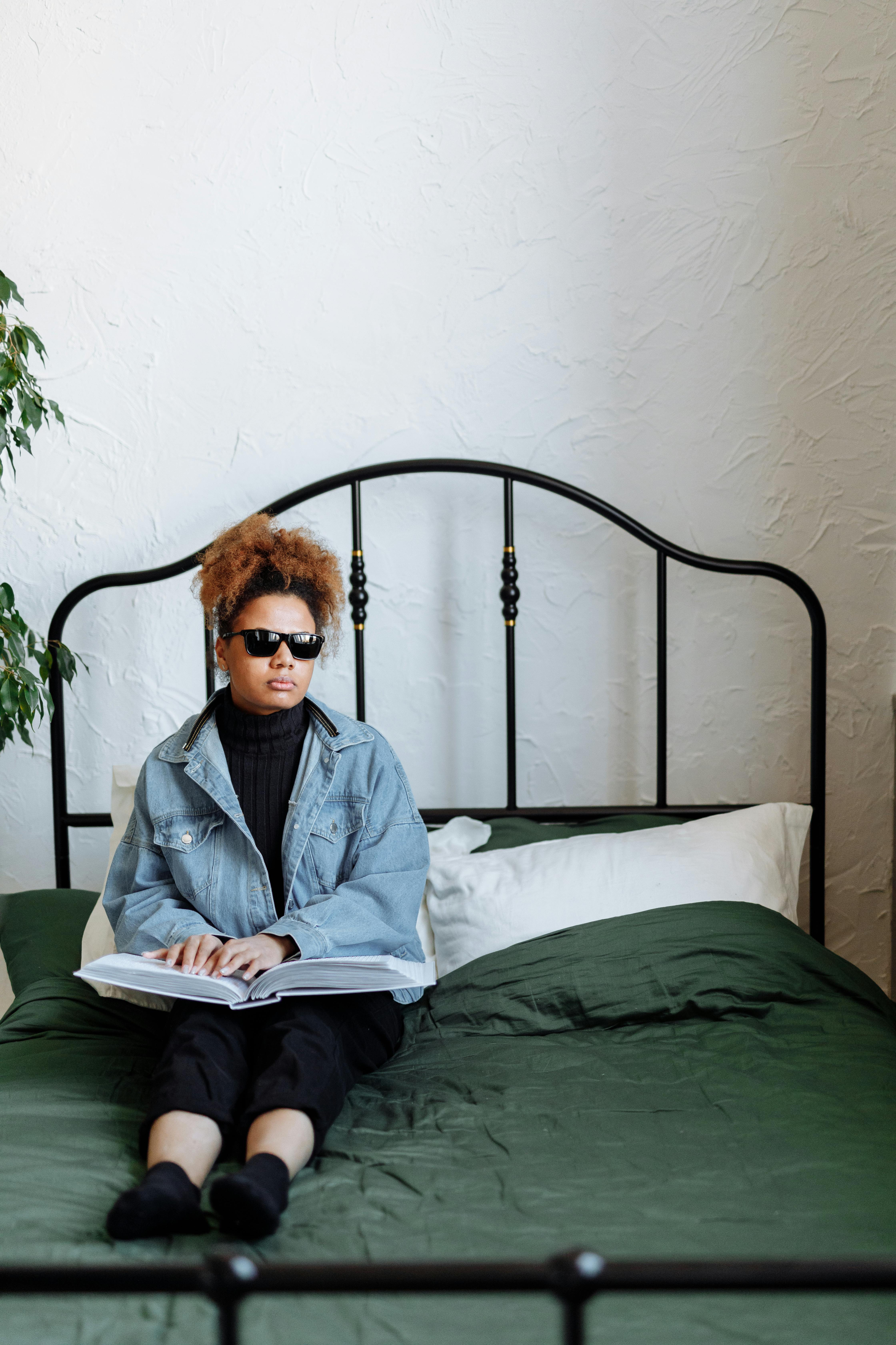 Adult woman in sunglasses reading a braille book on a bed in a stylish bedroom with a green bedspread.