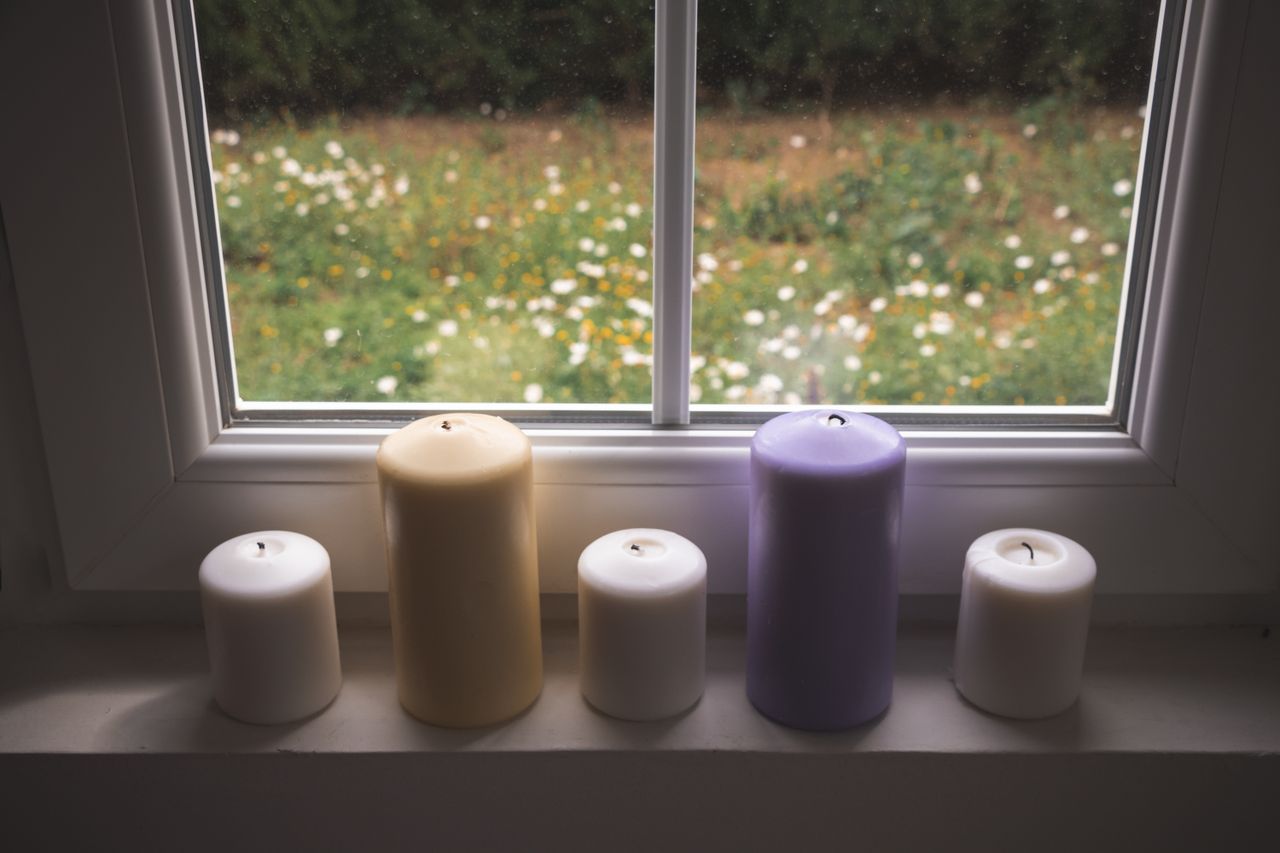 Five candles in white, cream and purple lined up on a windowsill with a garden view
