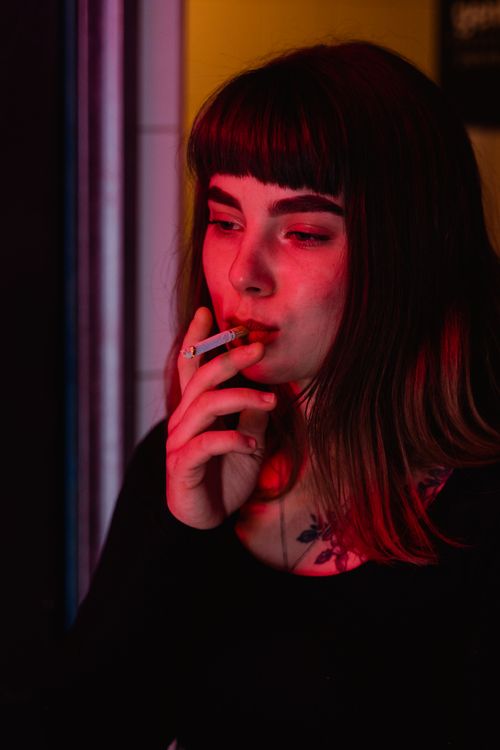 Free Portrait of a young woman with bangs smoking a cigarette under red lighting indoors. Stock Photo