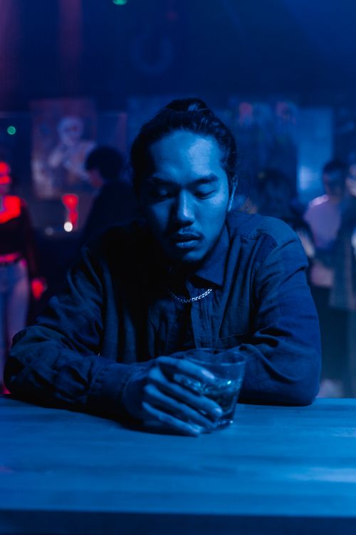 Free Man sitting alone at a bar holding a drink, surrounded by nightlife ambiance in a dimly lit room. Stock Photo