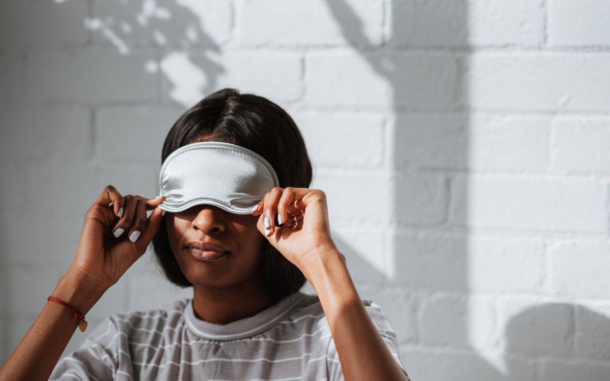 A woman wearing a grey striped shirt adjusts a sleeping mask in a bright indoor setting with shadows on the wall.