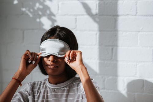 Free A woman wearing a grey striped shirt adjusts a sleeping mask in a bright indoor setting with shadows on the wall. Stock Photo