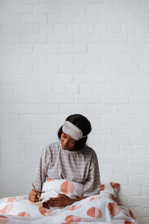 Free A Woman Sitting on the Bed while Writing on Her Notebook Stock Photo