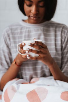 An African American woman enjoys a cozy morning with a ceramic mug, featuring stylish white manicure.