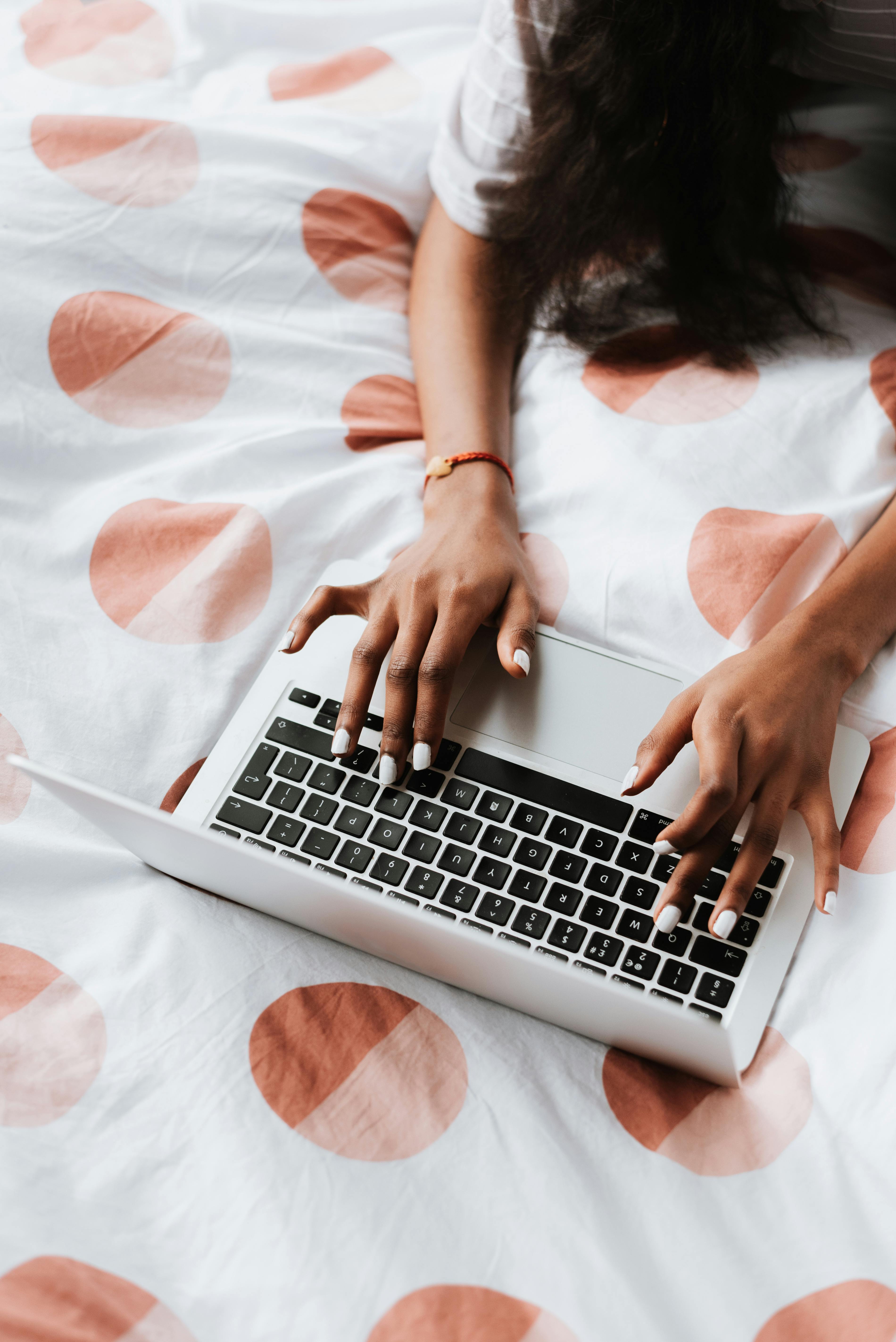 Top view of a woman with manicured nails typing on a laptop on a patterned bed.