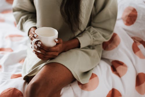 Free Crop anonymous ethnic female with cup of aqua sitting with crossed legs on bed blanket at home Stock Photo
