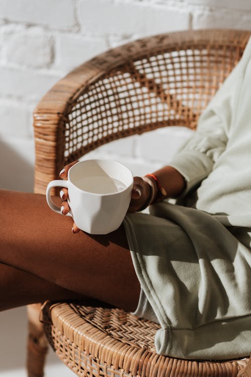 Free Crop anonymous black female with cup of aqua sitting with crossed legs in wicker armchair in sunlight Stock Photo