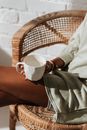 Crop anonymous black female with cup of aqua sitting with crossed legs in wicker armchair in sunlight
