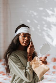 Young ethnic female cleaning cheek with cotton pad while holding mirror and fabric bag in bedroom