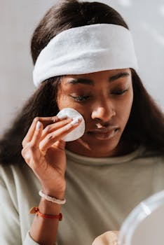 Young African American female in headband cleaning cheek with cotton pad while looking in mirror in sunlight