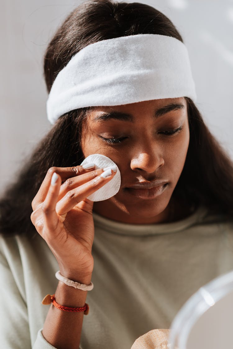 Black Woman Cleansing Face With Cotton Pad Against Mirror