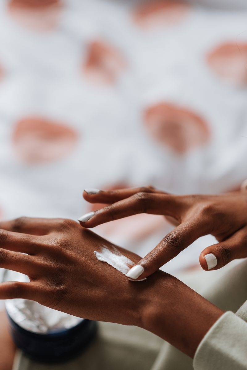 Close-up of a Black woman's hands as she smooths a stripe of white cream onto the back of one hand, an open jar of moisturizer beside her; everyday skin-care products like this are a common trigger of contact dermatitis.